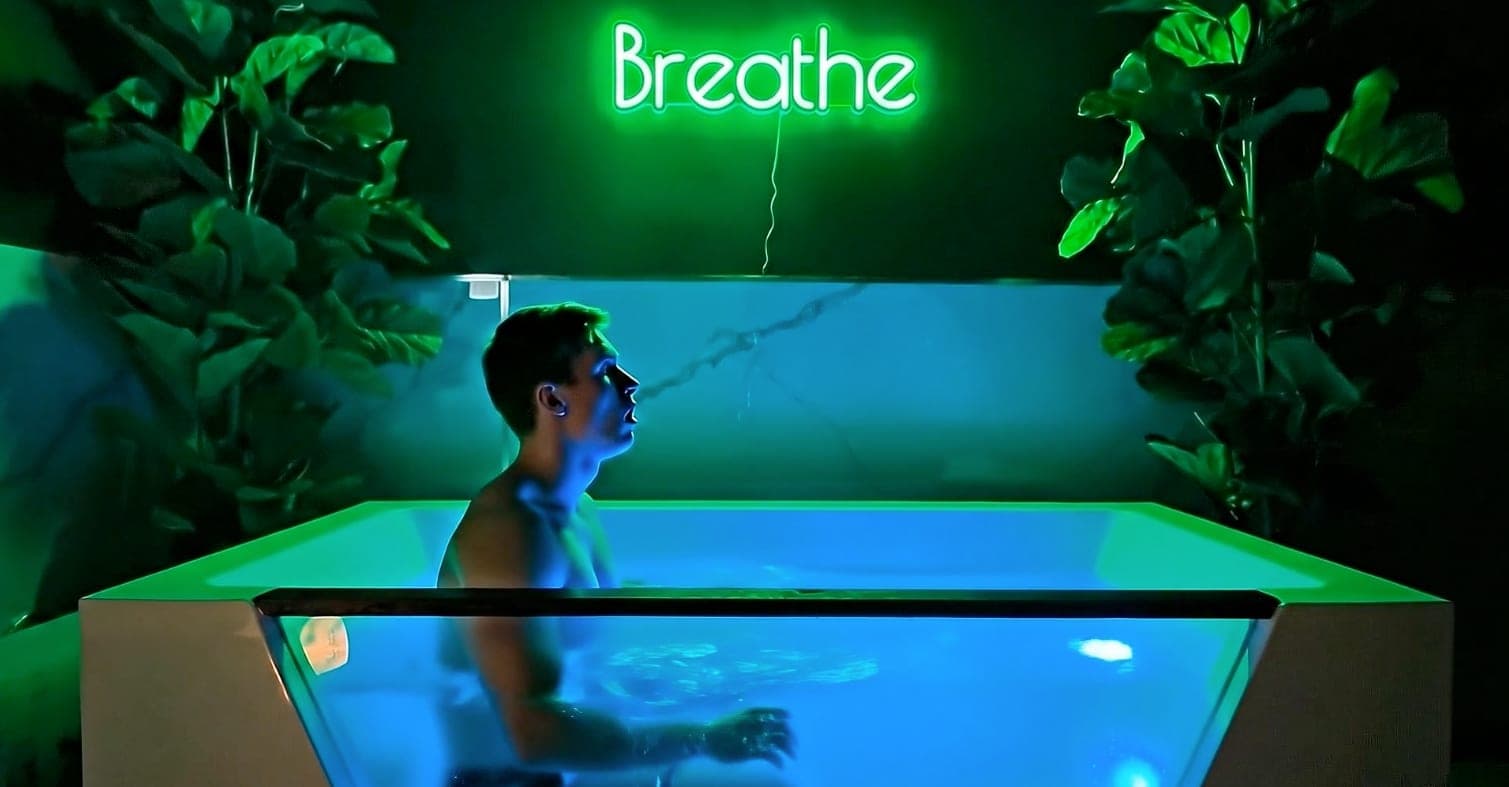 A guest sits chest-deep in a glass-sided cold plunge lit blue, under a green Breathe neon sign