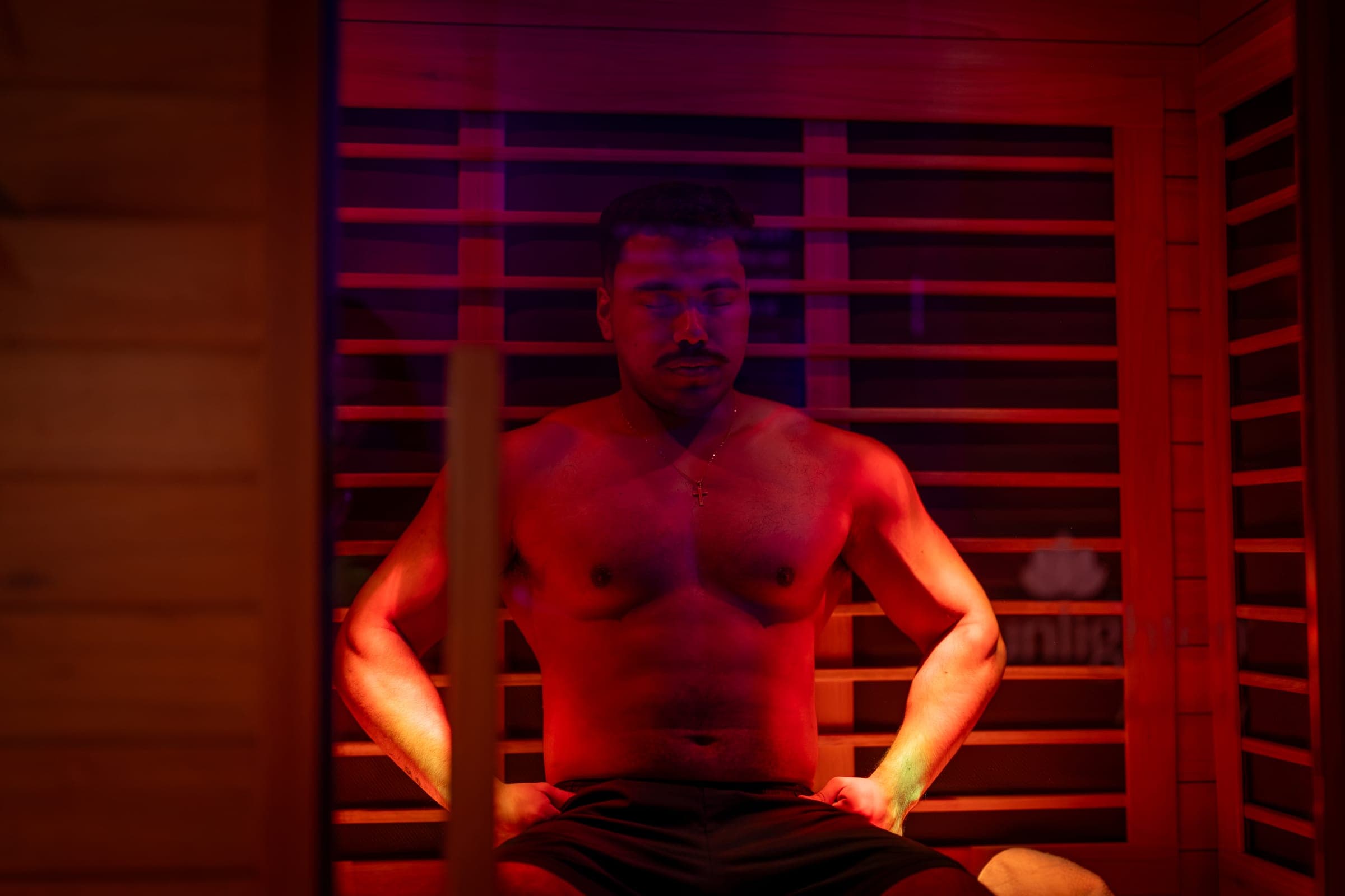 A guest sits forward on the infrared sauna bench in warm red light, mid-round